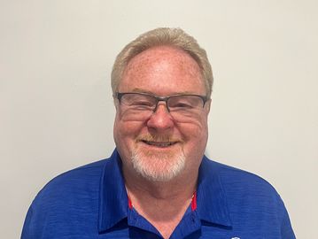 Smiling man with glasses and a blue polo shirt against a plain background.