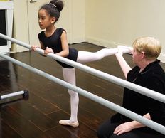 Ballet teacher helping young girl stretch on barre.