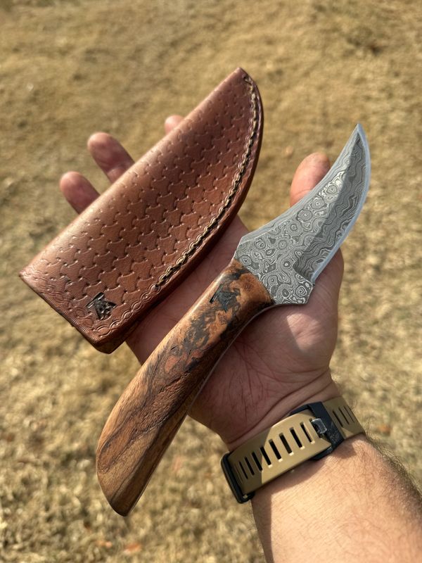 Hand holding a Damascus steel knife with a wooden handle and a brown leather sheath.