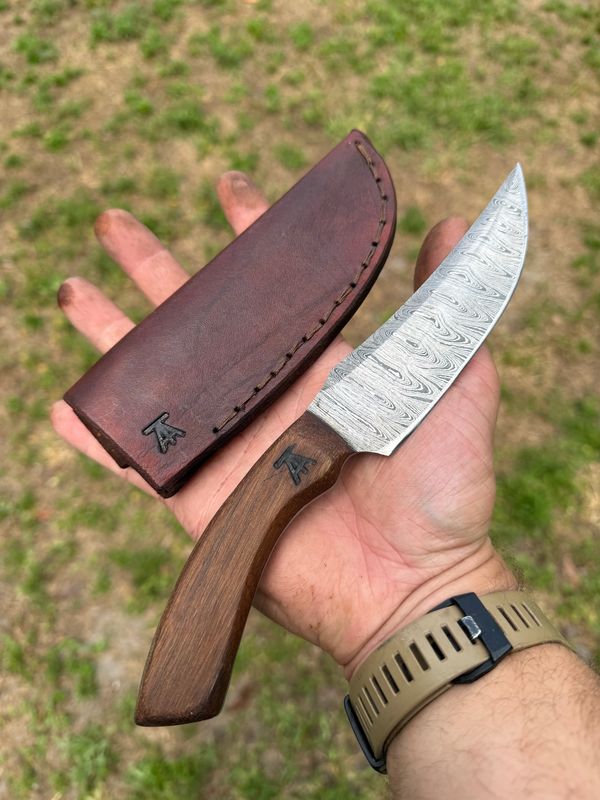 Hand holding a Damascus steel knife with wooden handle and brown leather sheath.