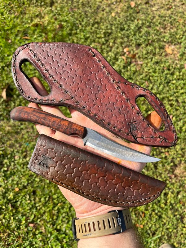 Hand holding a knife with two intricately stitched leather sheaths outdoors.