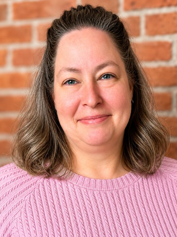 Smiling woman with shoulder-length hair in a pink sweater against a brick wall.