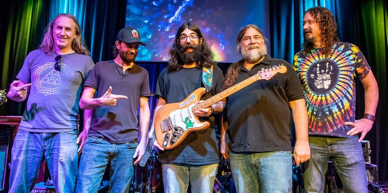 A group of five men posing on stage, one holding a guitar.