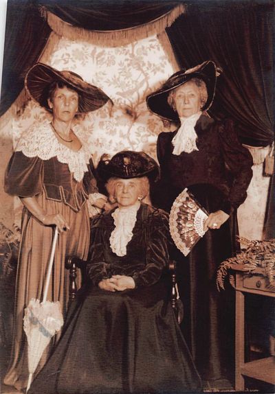 Three elderly women in vintage Victorian attire posing indoors with hats and accessories.
