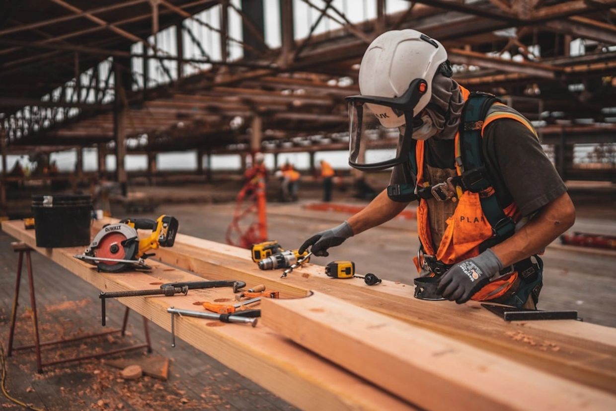 Construction worker wearing safety gear works on wooden beams in a large industrial space.