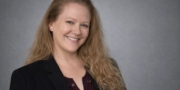 Smiling woman with long blonde hair in business attire against a gray background.
