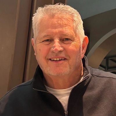 Smiling elderly man with white hair in a black jacket indoors.