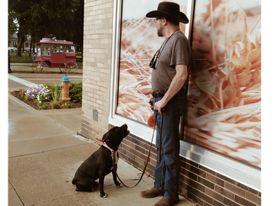 Man in cowboy hat with a black dog on a leash outside a building.