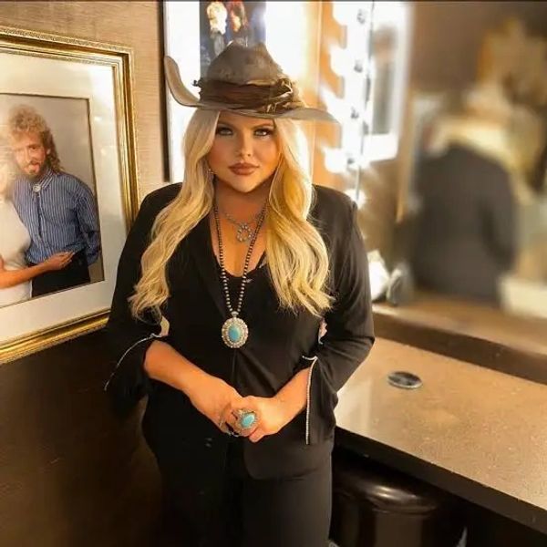 Woman with blonde hair wearing a hat and turquoise jewelry indoors.