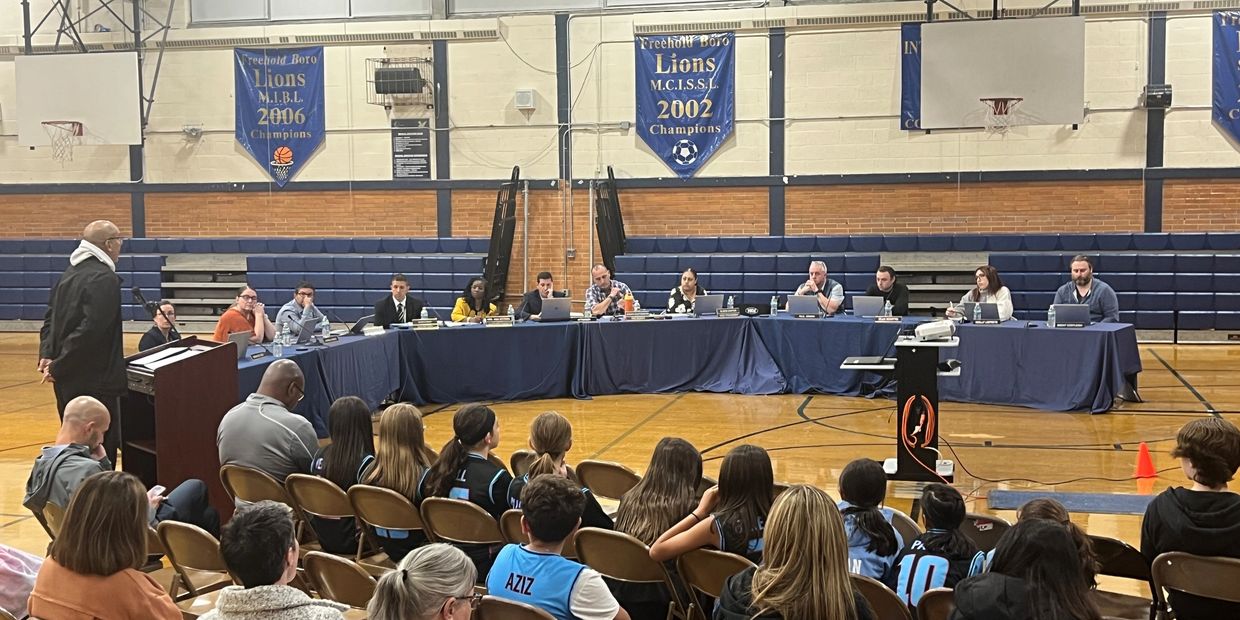A community meeting in a gymnasium with officials and attendees seated.