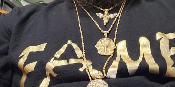 Person wearing gold necklaces over a black shirt with gold lettering.
