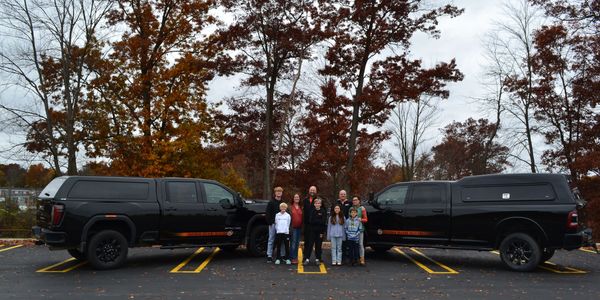 A group of people standing between two black trucks in a parking lot with autumn trees.