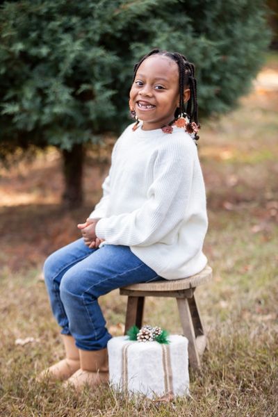 A smiling girl in a white sweater sitting on a stool outdoors.