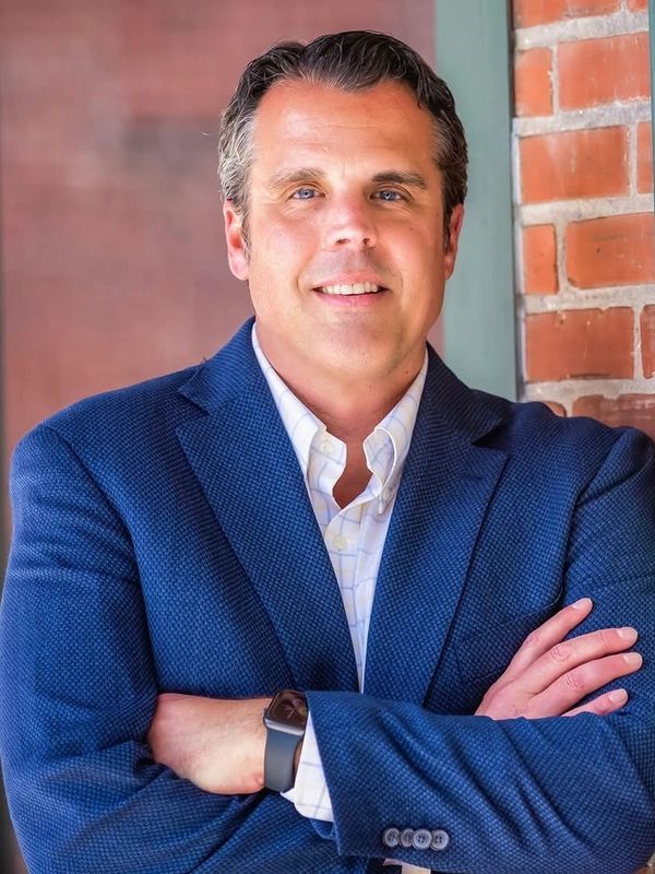 Confident man in a blue blazer standing with arms crossed against a brick wall.