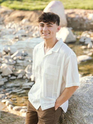 Teen boy smiling outdoors near a rocky stream, wearing a white shirt and brown pants.