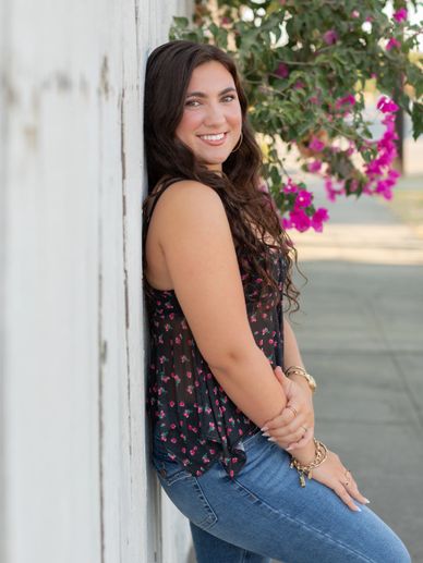 Smiling young woman leans against a white wall with flowers in the background.