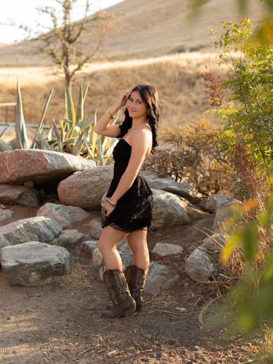 Woman in black dress and boots posing outdoors among rocks.