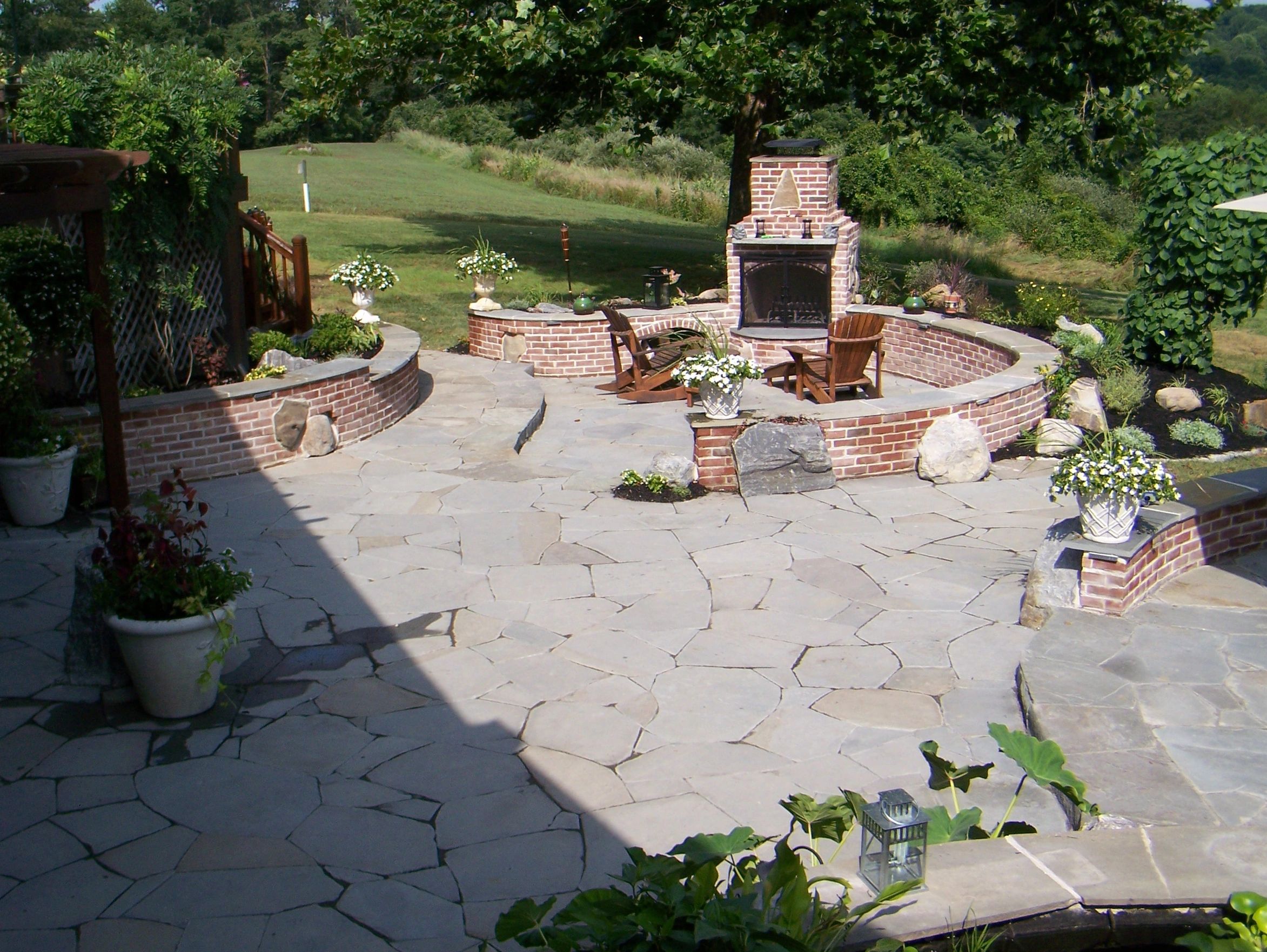 Charming patio with a brick fireplace, curved seating, stone flooring, and lush greenery