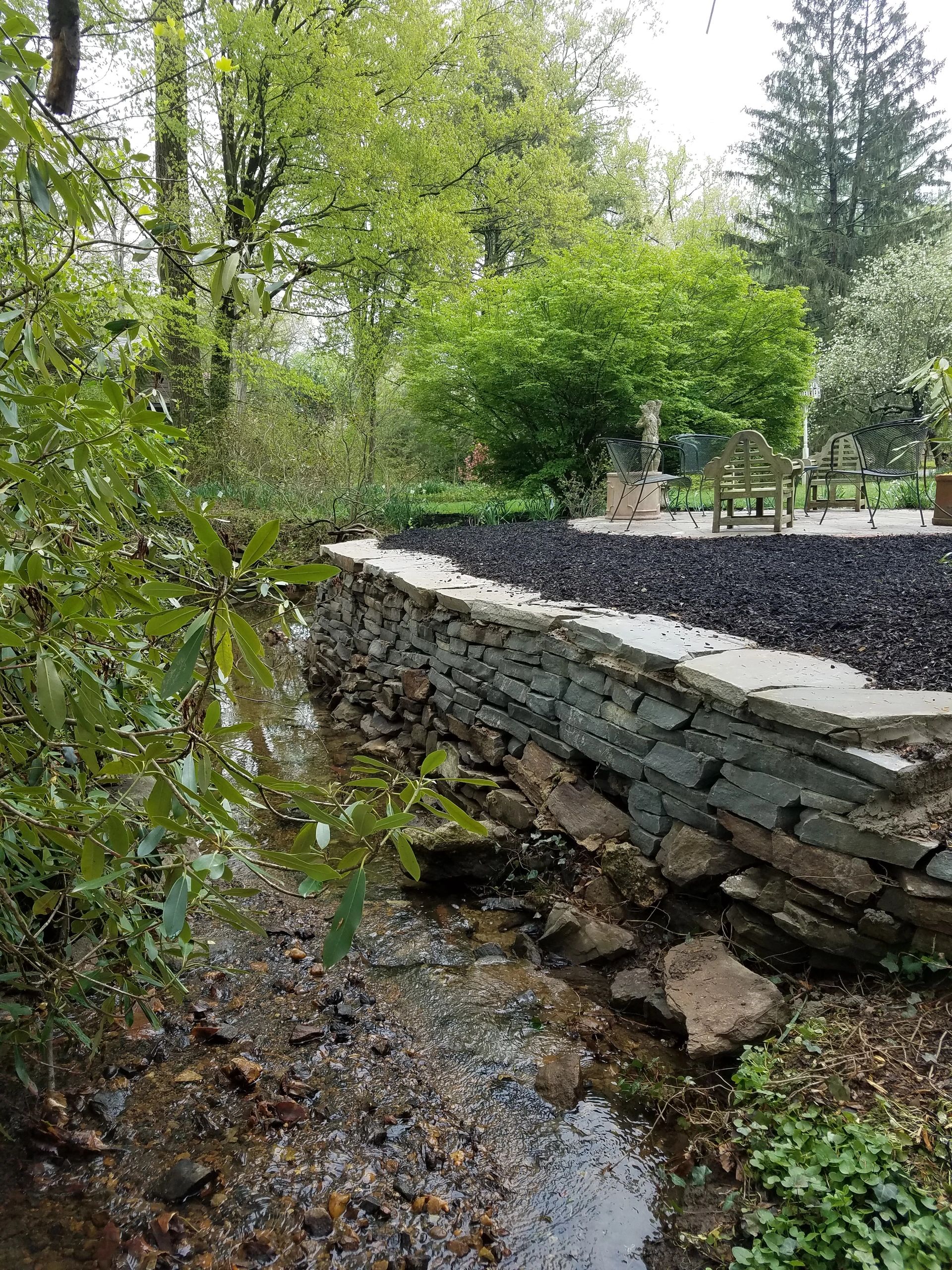 A stone retaining wall overlooking a small stream, surrounded by greenery, patio area with furniture