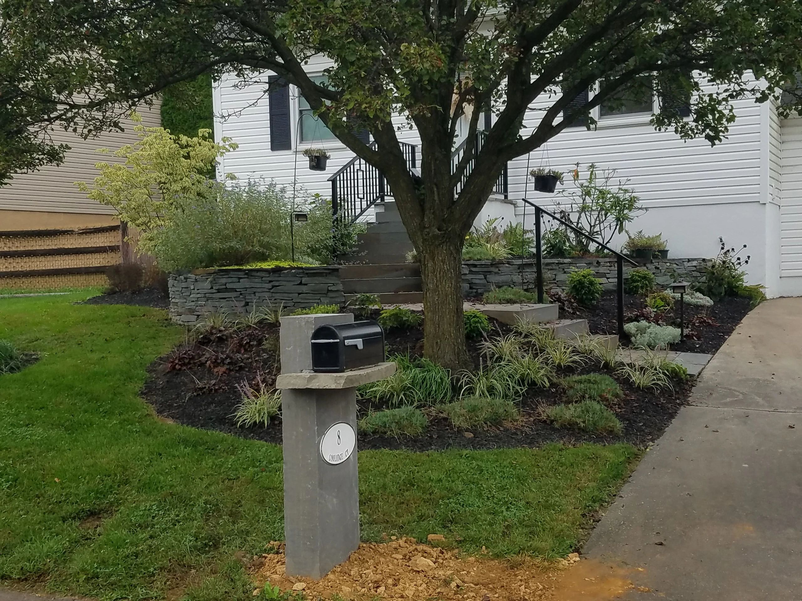 Front yard with a tree, landscaped garden beds, stone retaining walls, and a stone mailbox post.