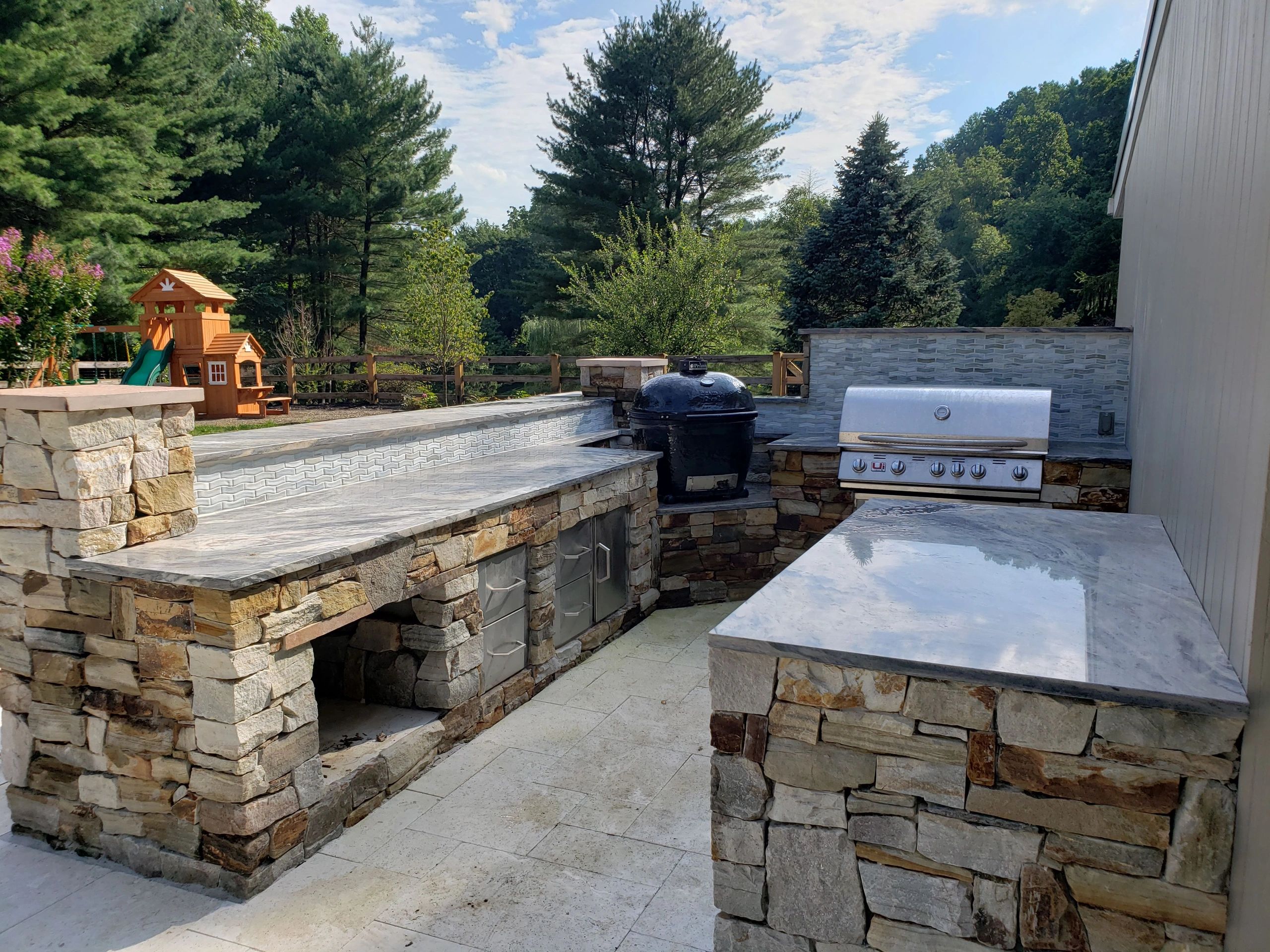 Outdoor kitchen with a stone counter, built-in grill, and a green backyard with a wooden playset.