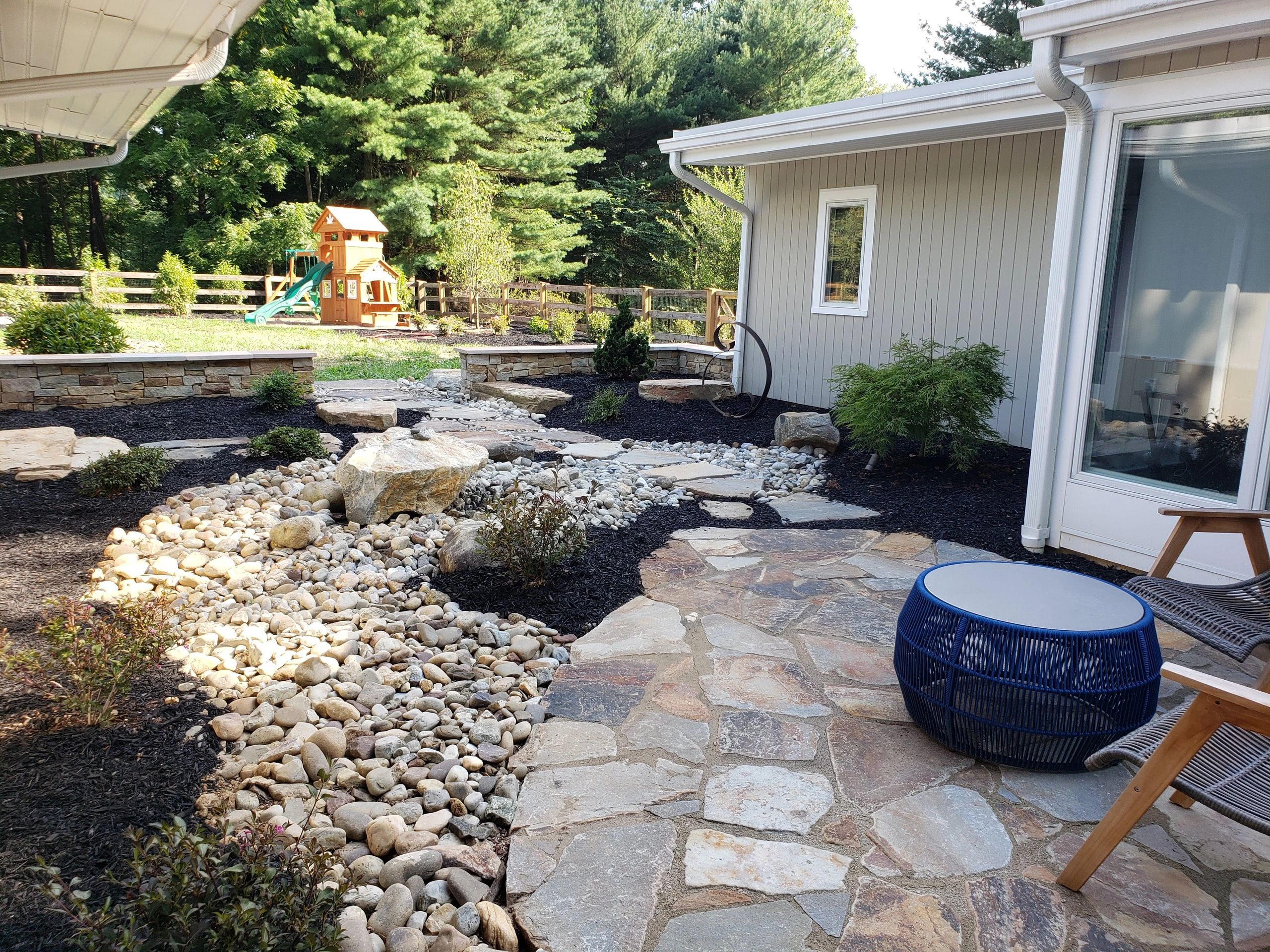 Backyard patio with stone pathways, a dry riverbed feature, and a wooden playset in the background.