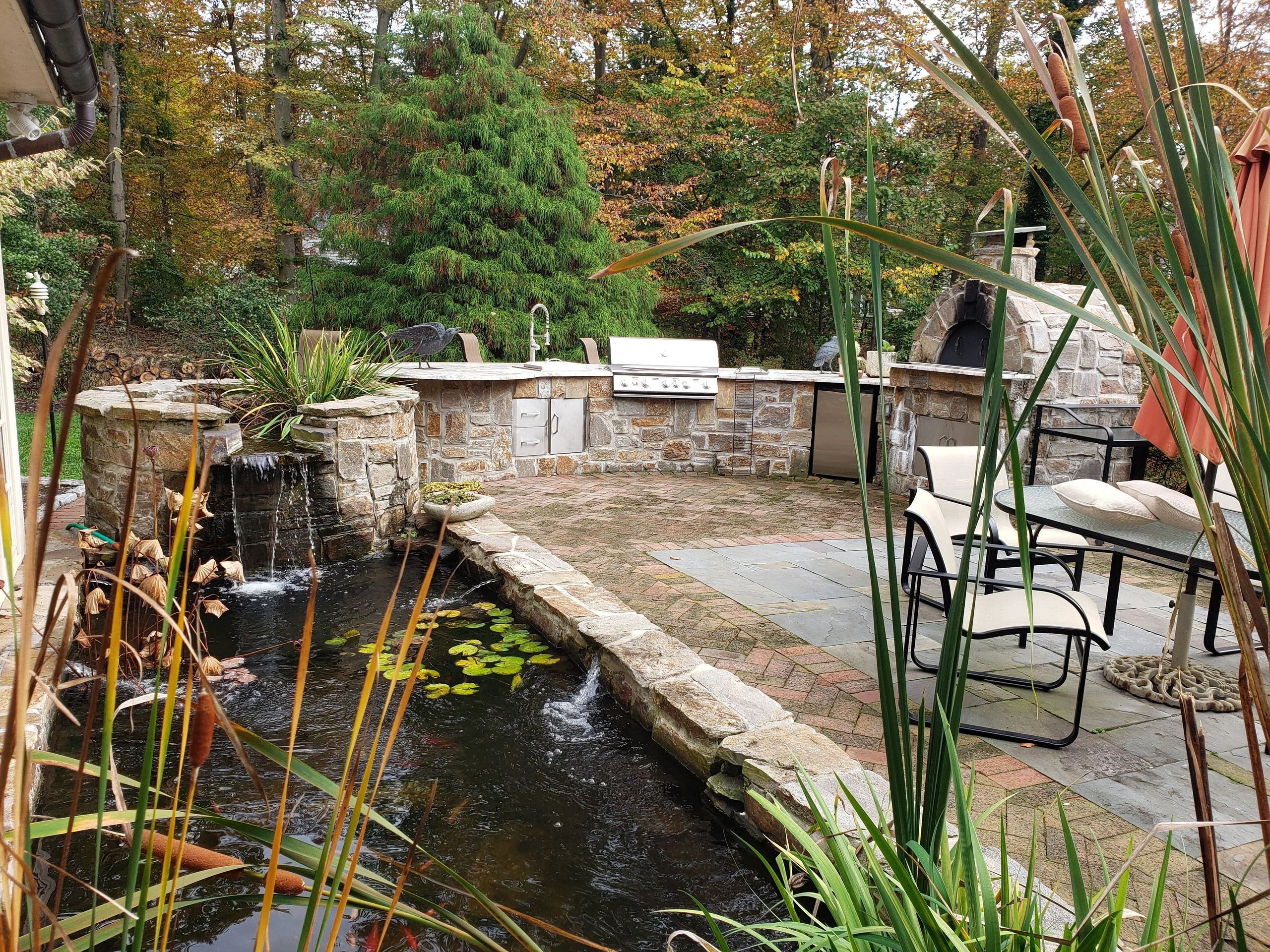 Outdoor kitchen with a stone countertop, grill, and pizza oven. a nearby koi pond with a waterfall.