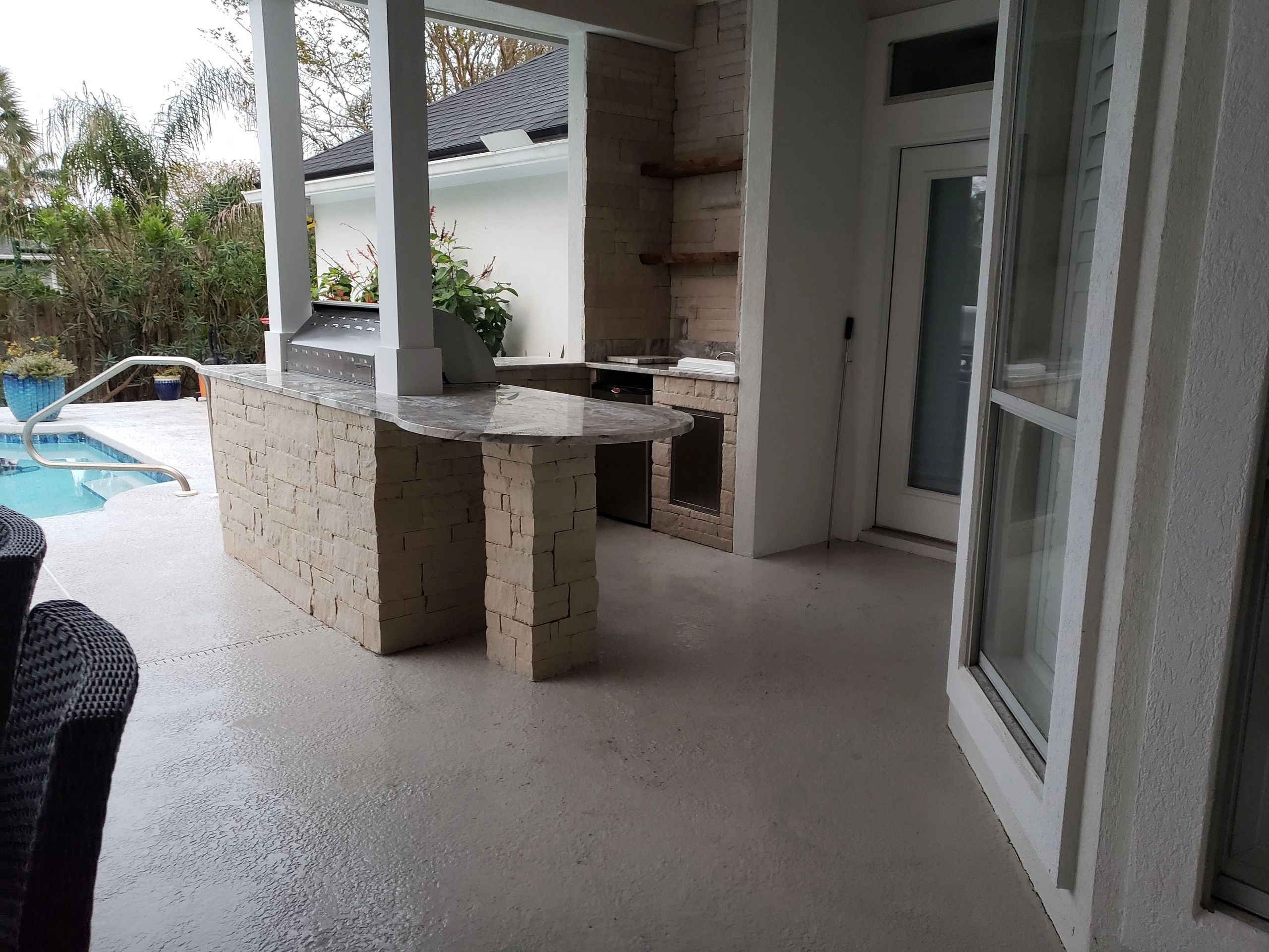 Covered outdoor kitchen with stone countertops, grill, sink and fridge. adjacent to a swimming pool.