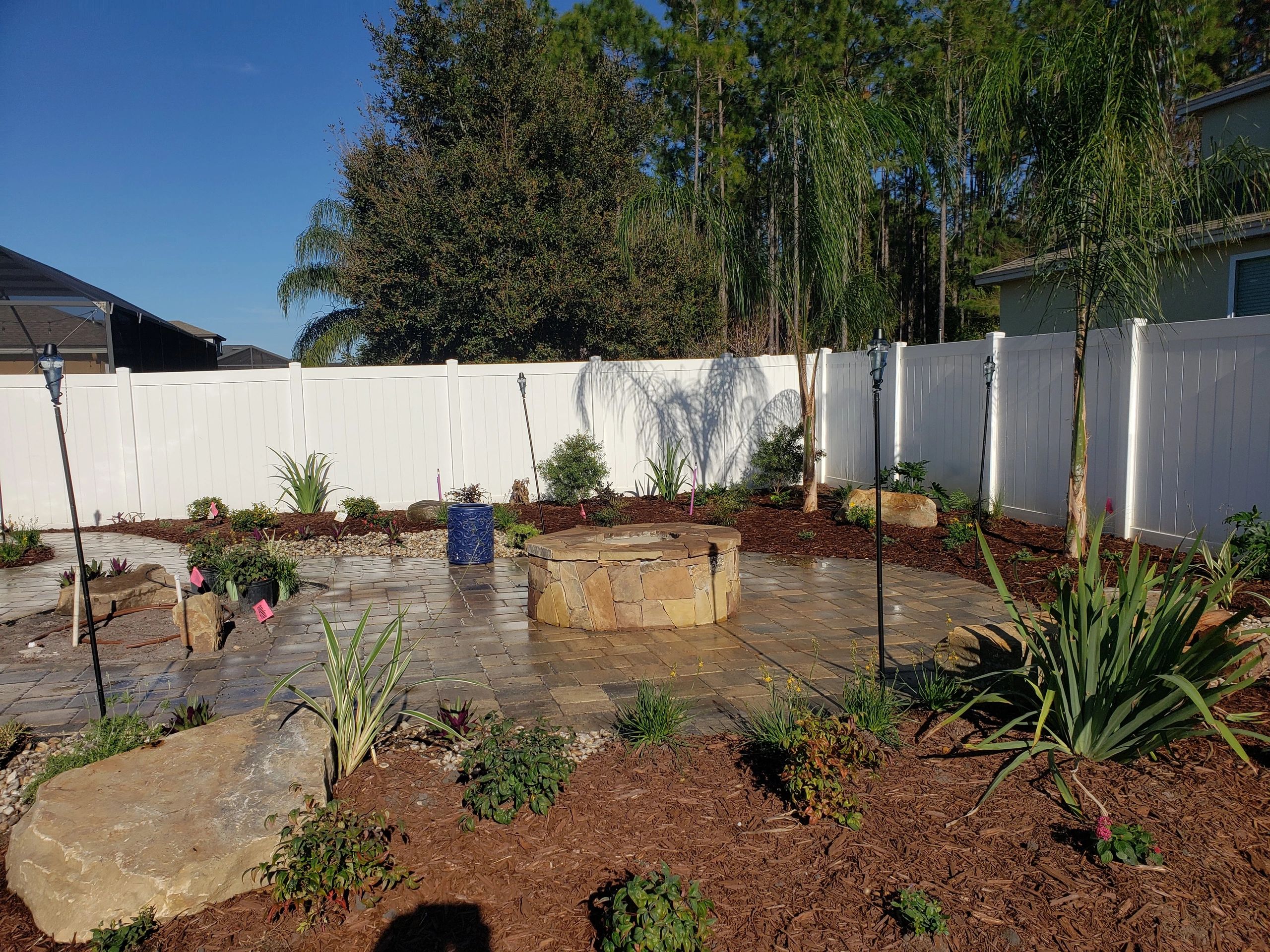 Backyard patio with a stone fire pit, surrounded by young plants, mulch, and a white fence. Nocatee.