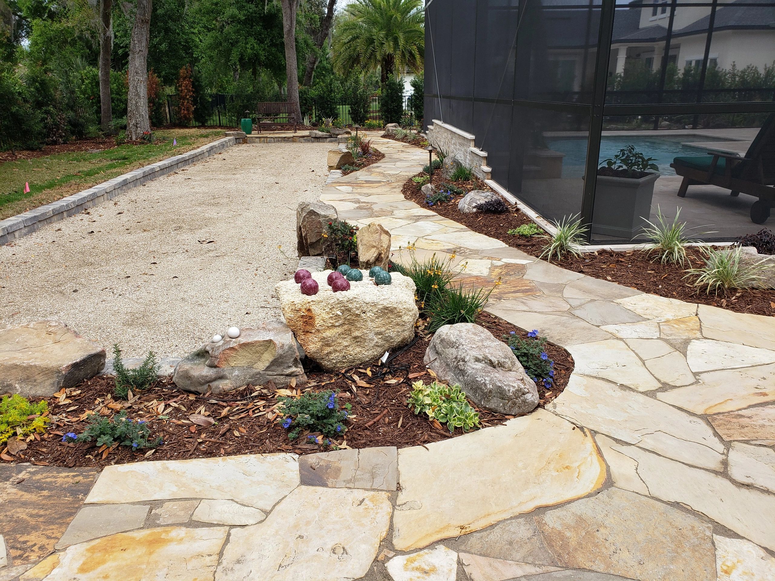 Backyard with a stone pathway, gravel bocce court, landscaped garden, and a screened pool area.