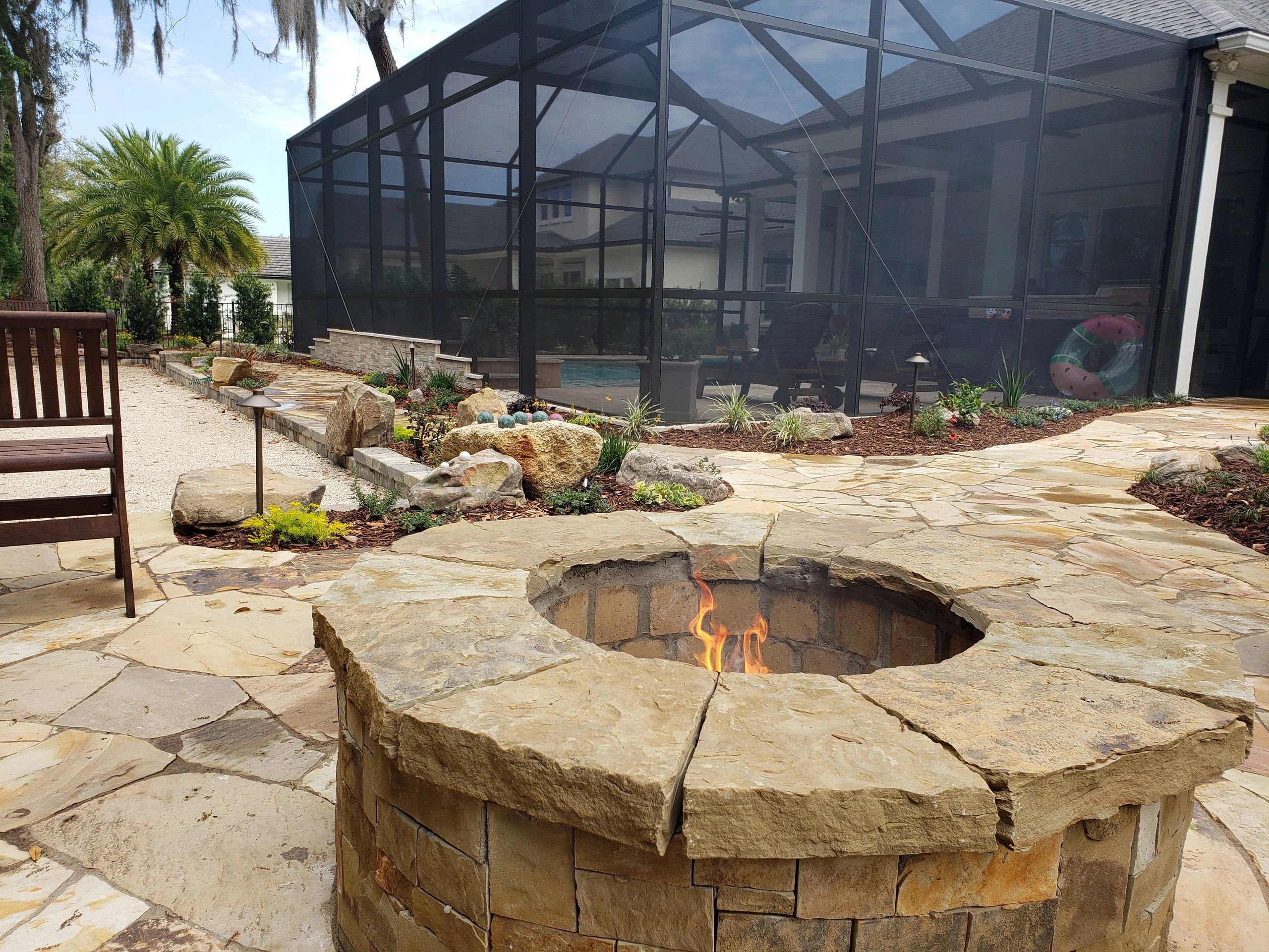 Stone fire pit on a patio with garden beds, a stone pathway, and a screened pool enclosure.