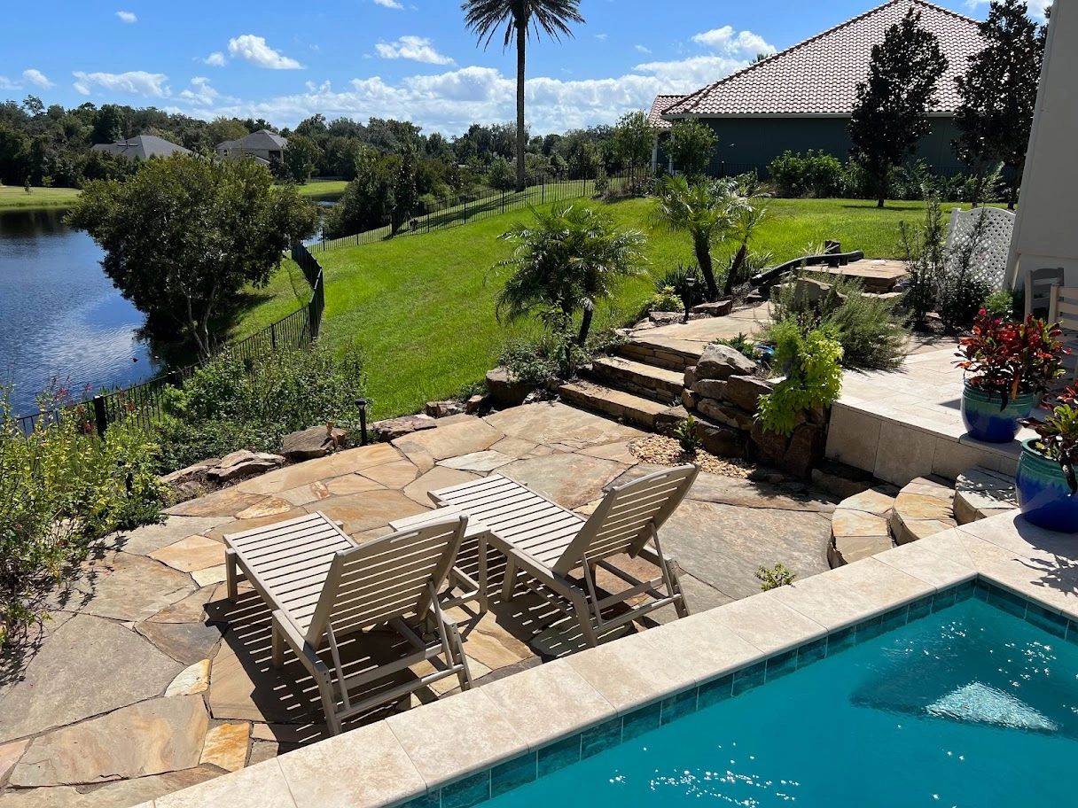 Poolside patio with stone flooring, lake view, lounge chairs, palm trees, and vibrant greenery.