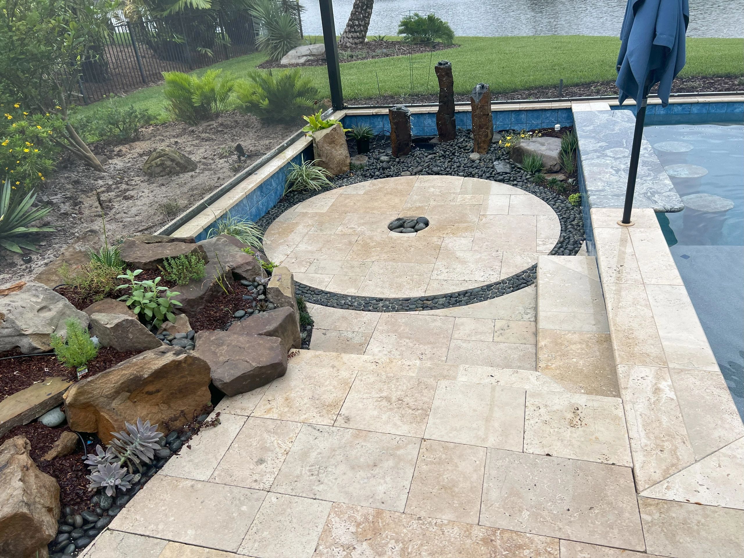 Poolside area with a circular stone pad feature surrounded by rocks, plants, a small water fountain.