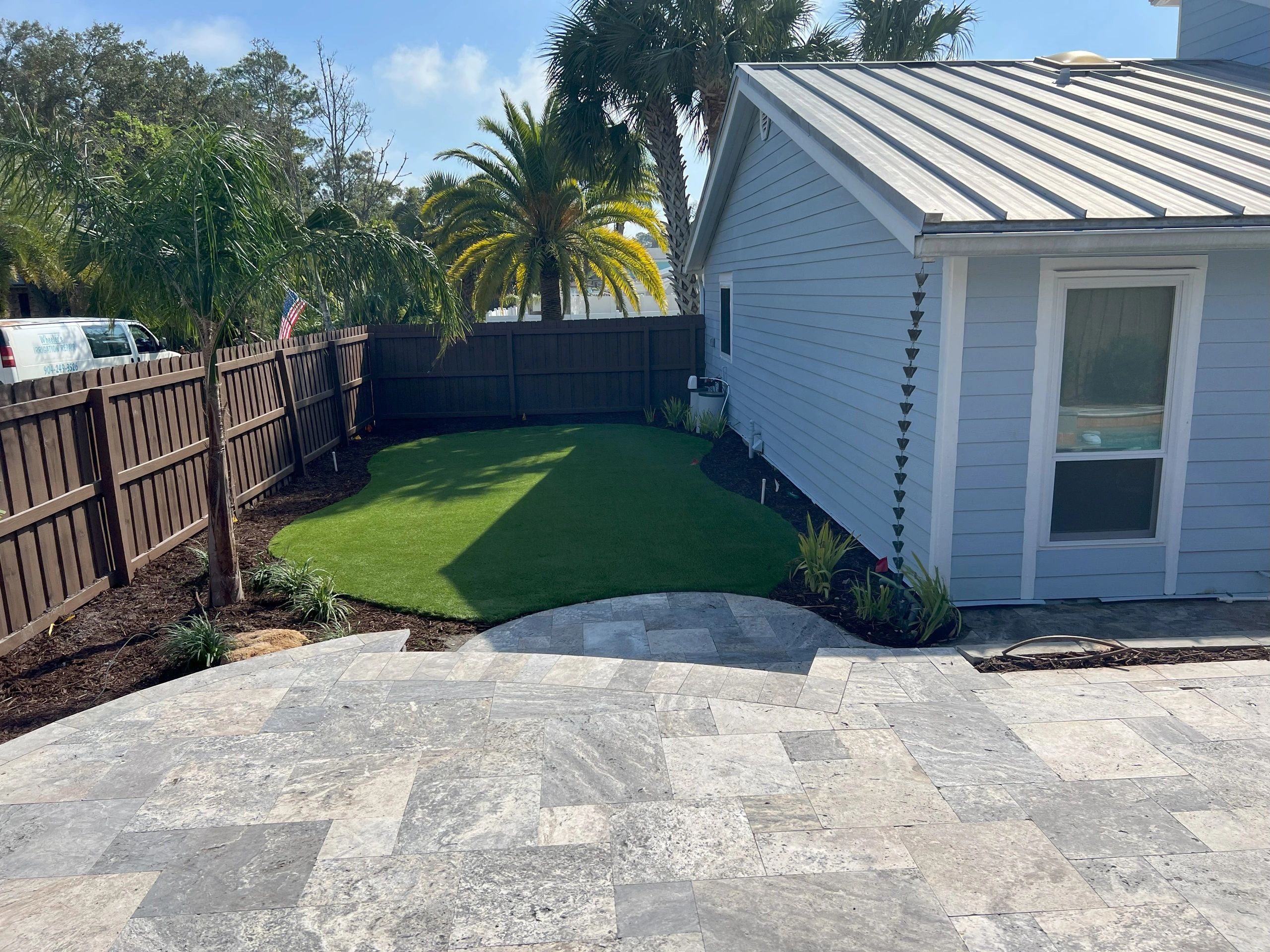 Backyard with a stone patio, artificial turf lawn, and palm trees, bordered by a wooden fence.