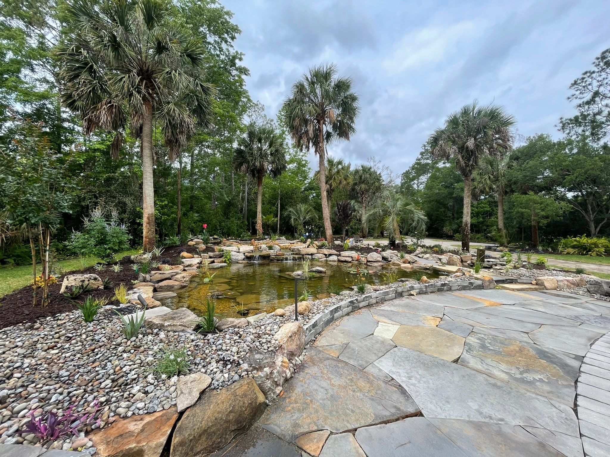 Pond with waterfall surrounded by palm trees and lush landscape. Boulders, river stone, flagstone.