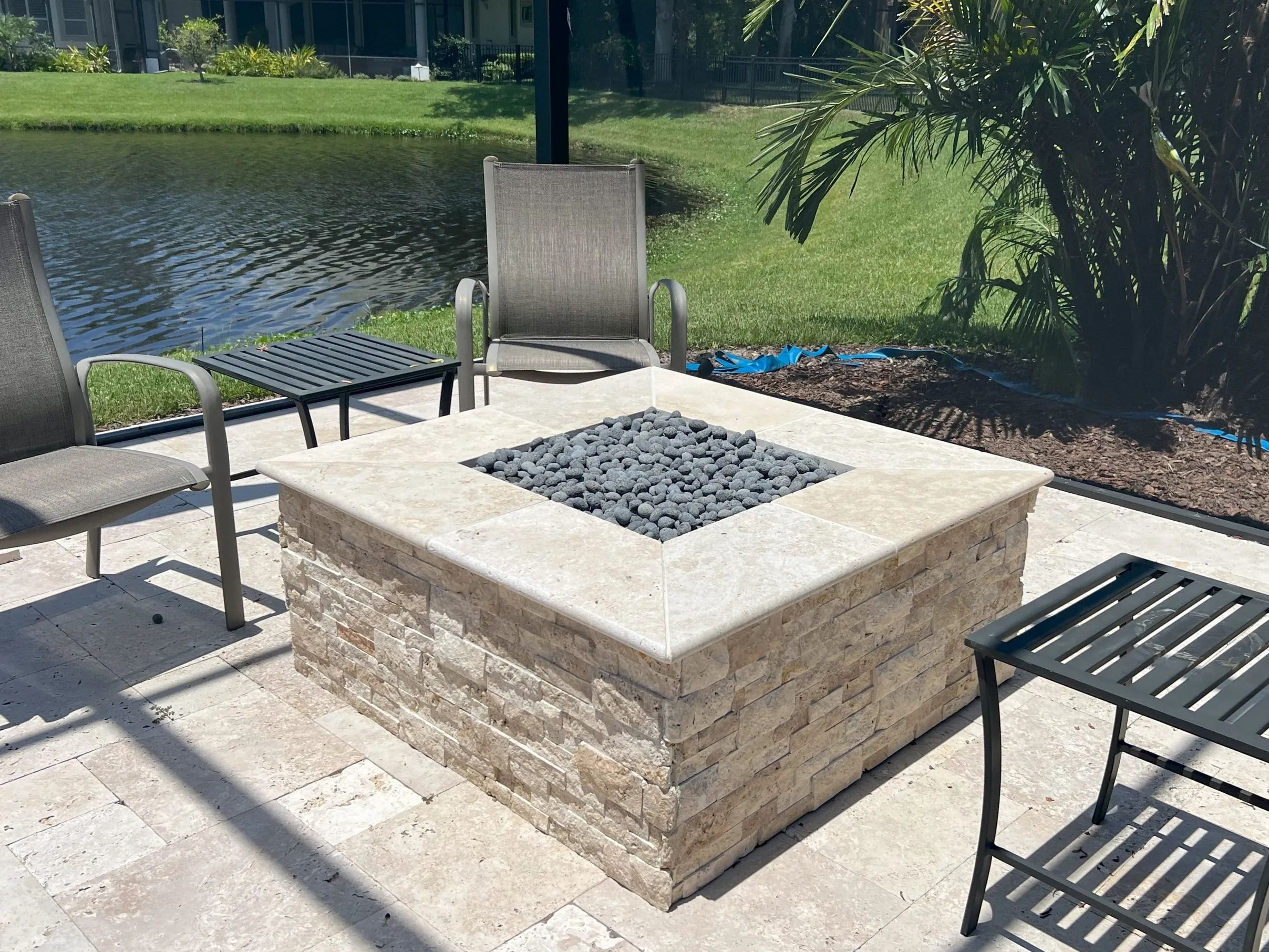 Outdoor patio with a square stone gas fire pit surrounded by chairs, overlooking a pond.