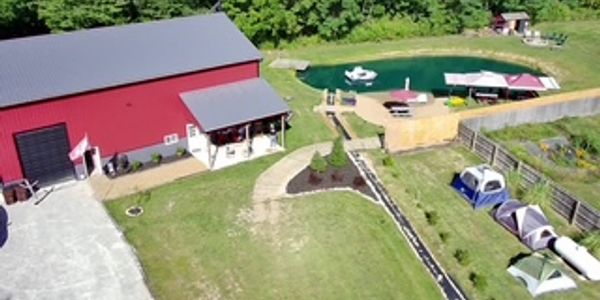 Red barn next to a pond with boats and tents in a green field.