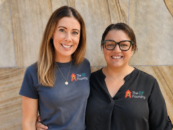 Two smiling women wearing The OT Foundry shirts stand against a stone wall.