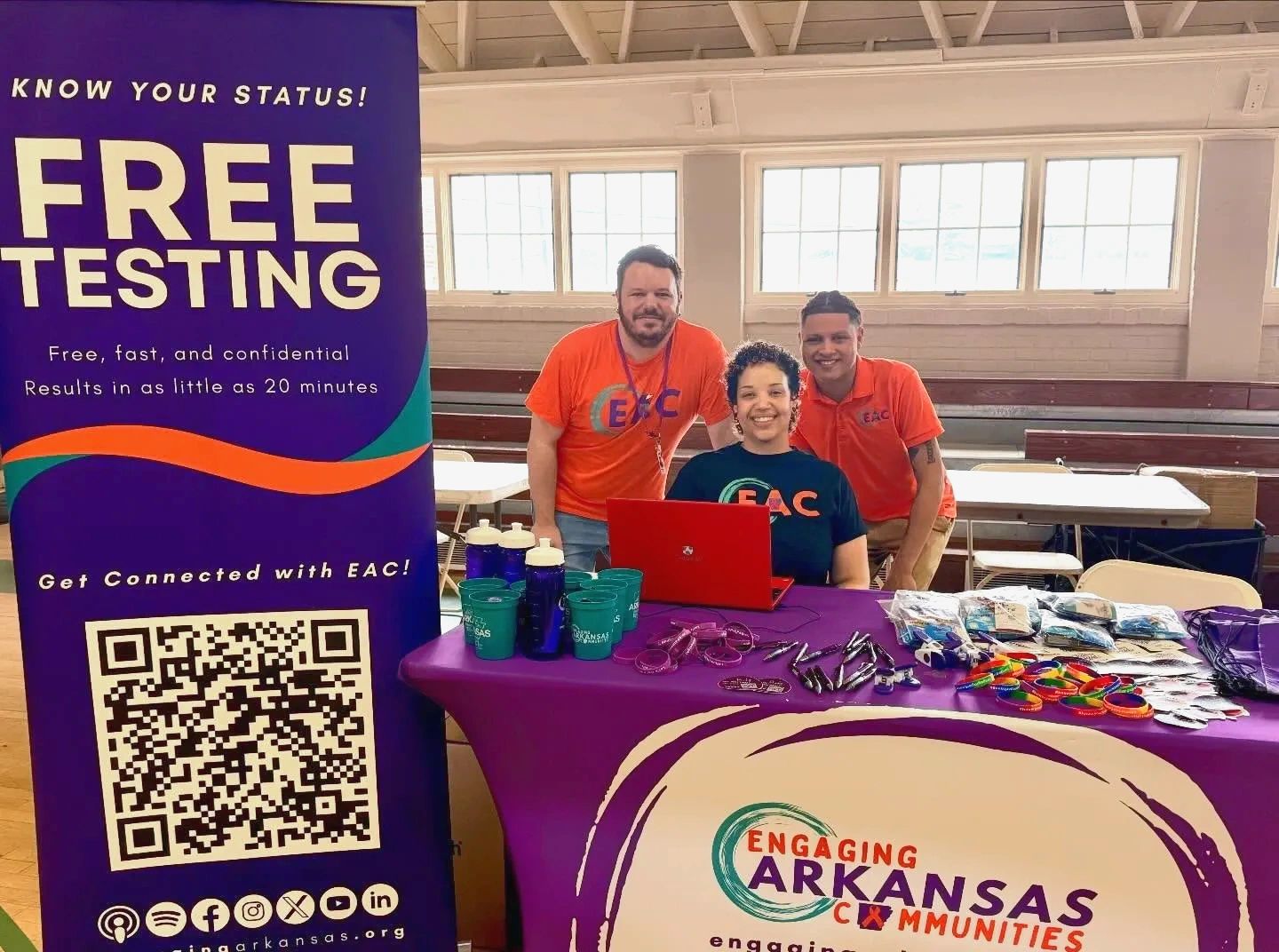 Three smiling people at a free testing event with informational table and sign.