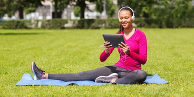 WorkoutStyles Fit woman exercising in a park using an ipad and head phones while sitting on an exercise mat
