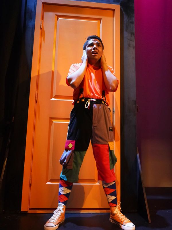 Person in colorful outfit covering ears in front of an orange door.