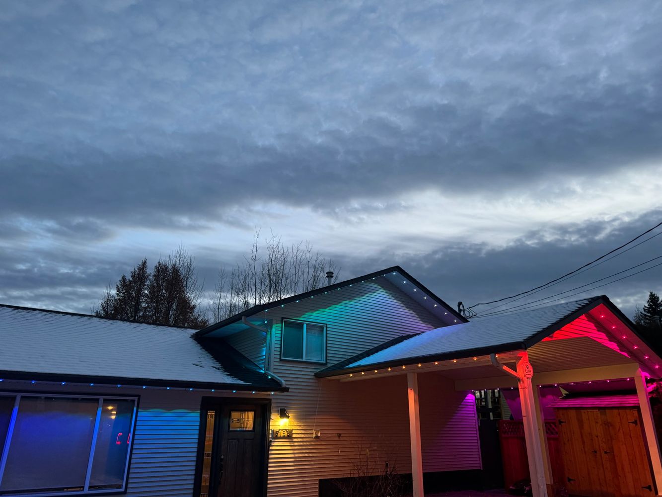 A house with snow-covered roof illuminated by colorful lights under a cloudy sky.