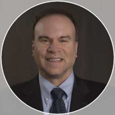Professional headshot of a smiling man in a suit and tie against a dark background.