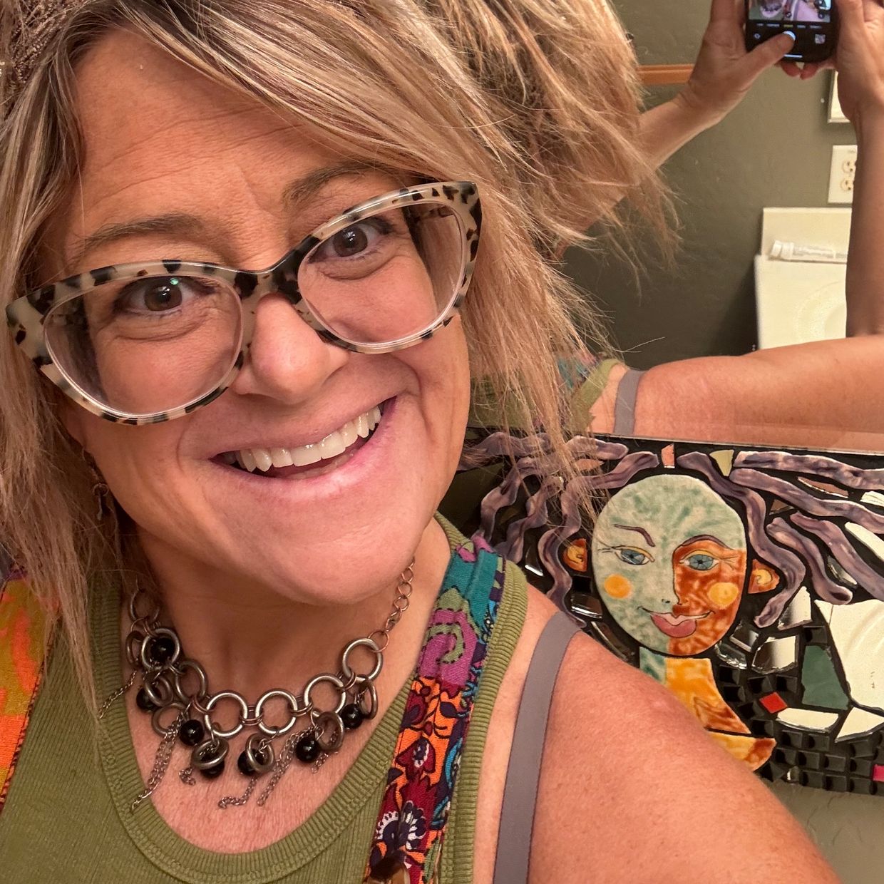 Smiling woman with glasses taking a colorful selfie in front of a mosaic artwork.