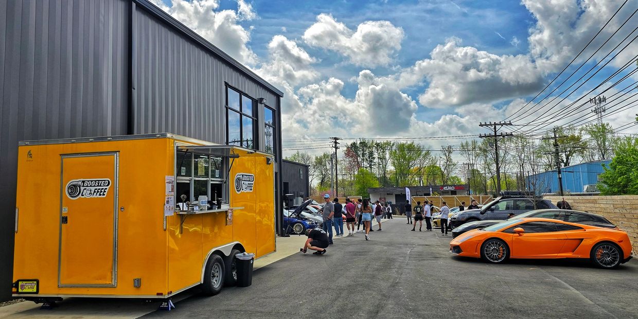 A bright yellow coffee trailer and an orange sports car at an outdoor event with people and blue sky.