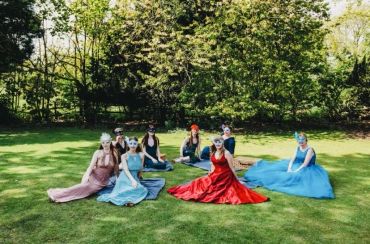 Seven women in colorful dresses and masks sitting on grass in a garden.