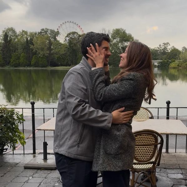 Pareja abrazándose cerca de un lago con una noria al fondo.