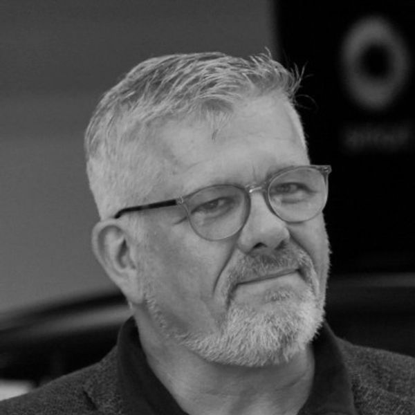 A mature man with gray hair and glasses, wearing a dark jacket.