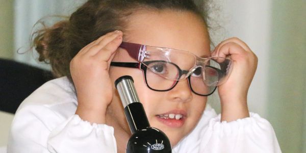 Young girl wearing safety glasses and looking through a microscope.