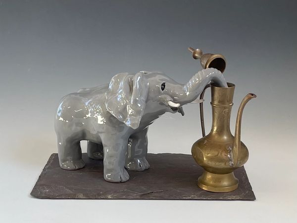 a grey elephant with his trunk in a brass ewer all mounted on a piece of slate